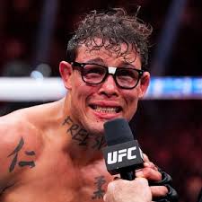 Brandon Moreno, lutador de MMA, com óculos e microfone da UFC, expressando emoção após luta.