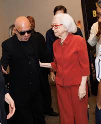 Homem careca de óculos escuros e mulher idosa de terno vermelho em evento.