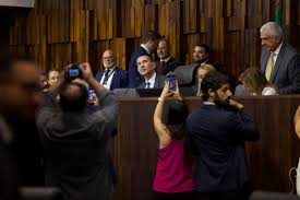 Homens e mulheres em um plenário, alguns tiram fotos com celulares. Possível evento político ou legislativo.