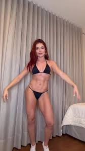 Mulher ruiva com abdômen definido posa usando biquíni preto em frente a cortinas.