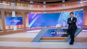 Apresentador do SBT Brasil em estúdio com telas exibindo o logo do canal e globo terrestre.