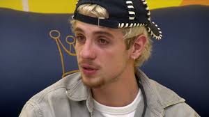 Gabriel Santana, participante do BBB 23, usa boné e jaqueta em close-up. Ao fundo, logo do programa com uma coroa.