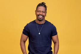 Homem negro sorrindo com cabelo preso em coque e camiseta azul escura, em fundo amarelo.