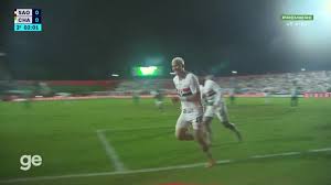 Jogador comemora gol em partida de futebol, com placar indicando São Paulo contra Chapecoense. Campo iluminado à noite.