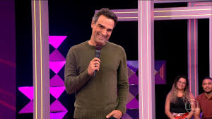 Tadeu Schmidt, apresentador do BBB, sorri segurando um microfone em um palco com luzes coloridas.