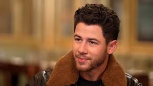 Nick Jonas sorri em close-up, usando jaqueta com gola de pele marrom. O cantor e ator está em destaque.