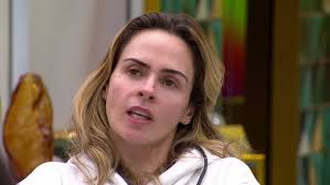 A atriz Deborah Secco em close-up, com cabelo loiro ondulado e maquiagem leve, falando em um ambiente interno.