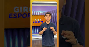 Jovem apresentador em estúdio com logo 'Giro Esporte' ao fundo, em programa de notícias esportivas.