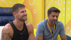 Dois homens sorriem em um ambiente com decoração amarela e azul, possivelmente em um reality show.
