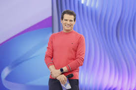 Apresentador Rodrigo Faro sorri para a câmera em um estúdio de TV com fundo azul e roxo.