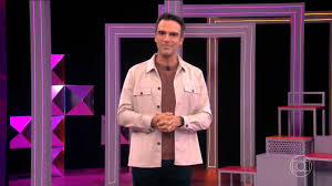 Tadeu Schmidt apresenta o Big Brother Brasil em cenário moderno com luzes roxas e detalhes geométricos.