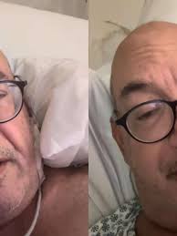 Homem careca com óculos de armação preta em leito hospitalar, com expressão preocupada.