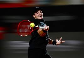 Tenista joga partida com raquete vermelha e bola amarela em quadra com fundo desfocado.