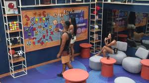 Participantes do BBB 23 interagem em sala com painel temático e móveis coloridos. Ambiente interno com espelhos e prateleira…