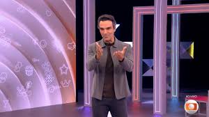 Tadeu Schmidt, apresentador do BBB 23, em um estúdio com iluminação neon e elementos gráficos de festa.