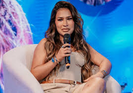 Simone Mendes sentada em uma poltrona branca, segurando um microfone, com um fundo azul e rosa.