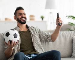 Image of person holding a soccer ball while sitting on a couch watching a game on TV